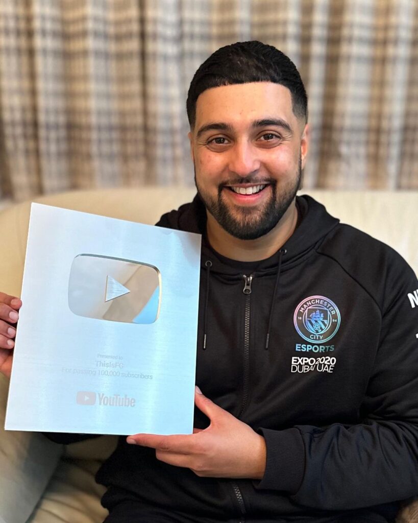 A beaming man holds a YouTube Silver Play Button award, recognizing his channel 'ThisIsFG' for surpassing 100,000 subscribers, dressed in a black hoodie with the Manchester City Esports logo.